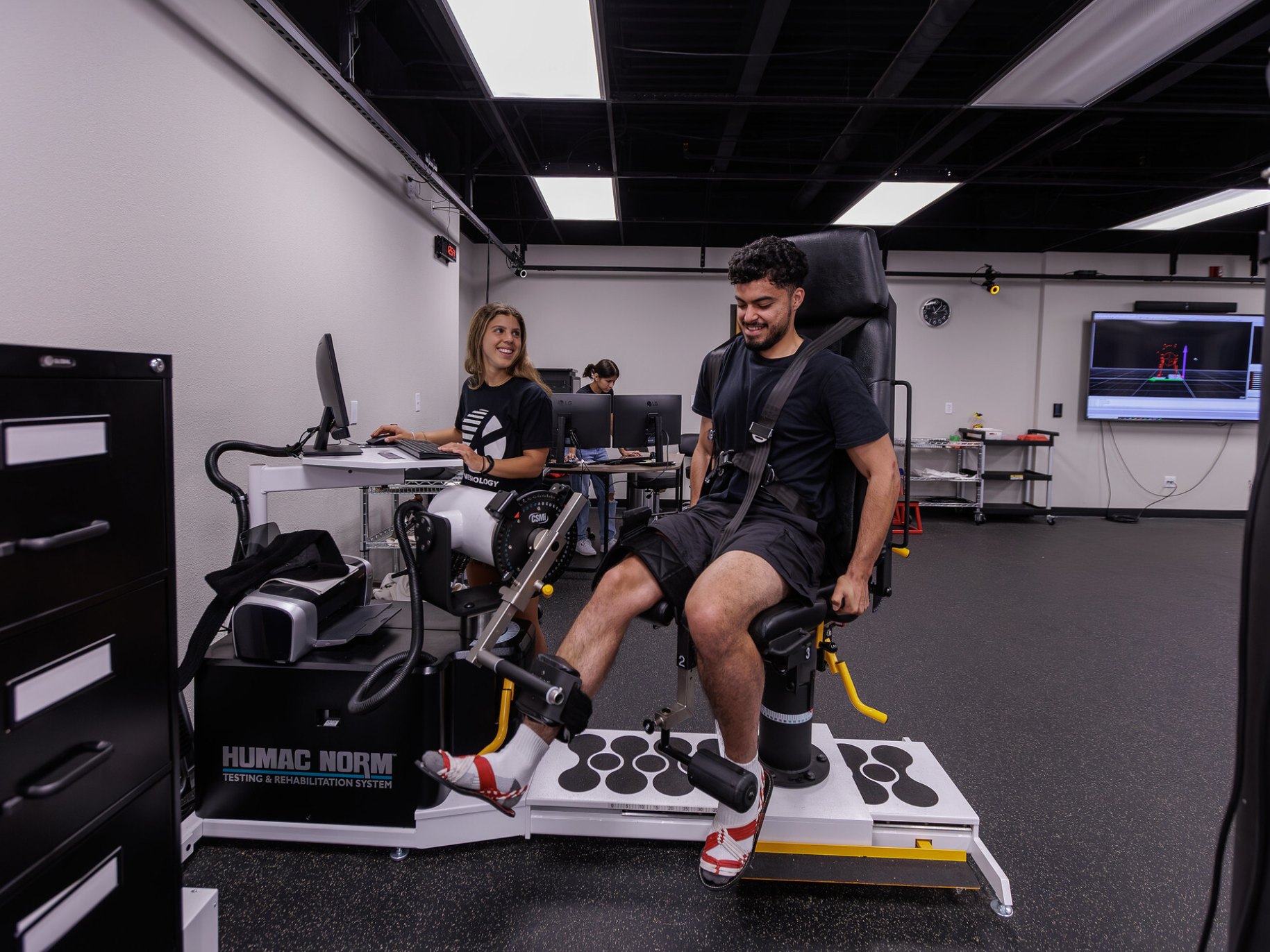 A man performs a leg exercise on a HUMAC NORM rehabilitation machine while a woman monitors him on a computer in a lab
