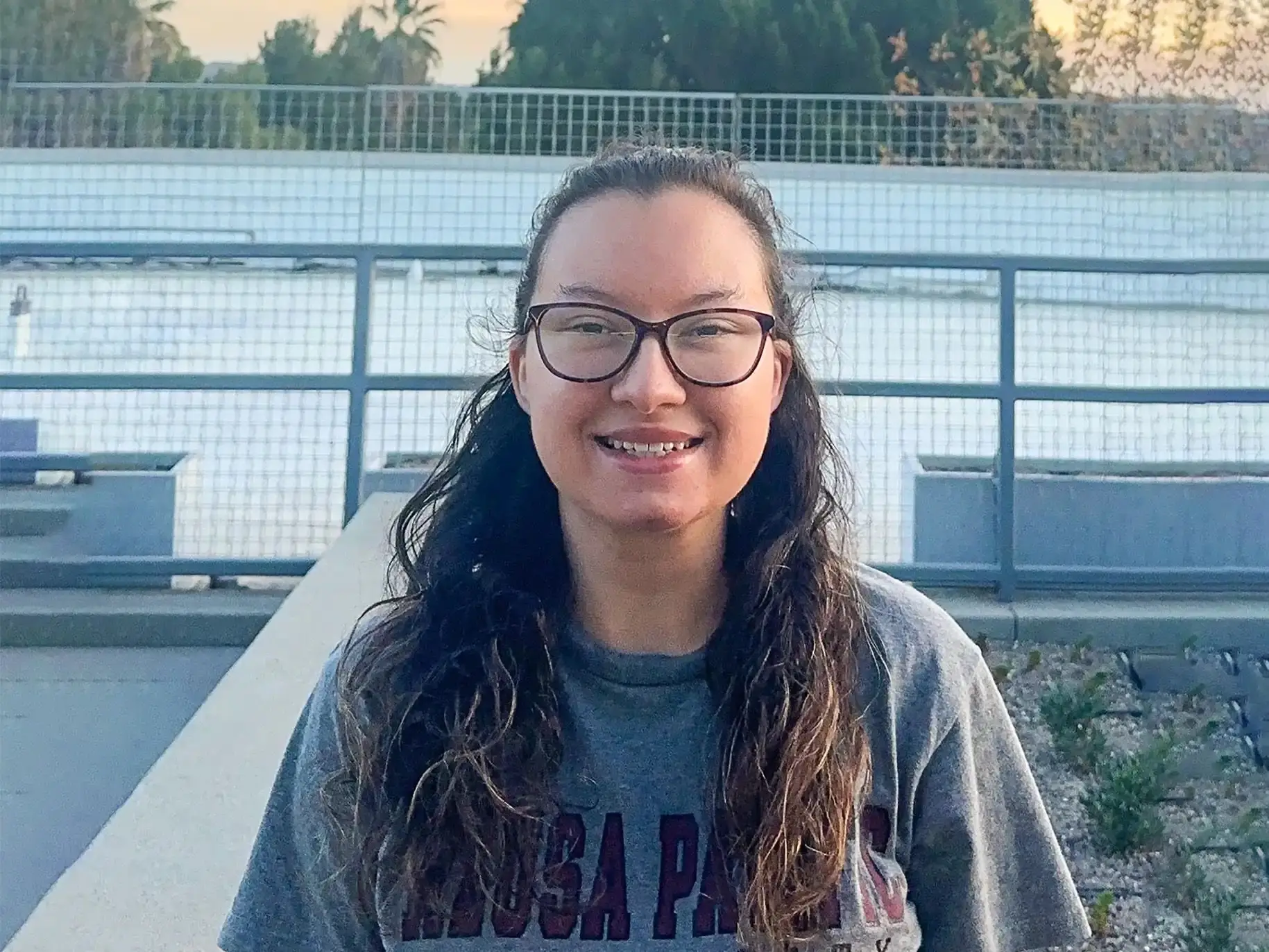 Emma smiles outside a field at APU, wearing a university t-shirt