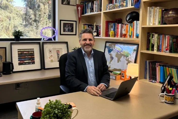 Ismael smiles at his desk, his office filled with a hodgepodge of items, such as a map of the world, colorful books, and soccer memorabilia. 