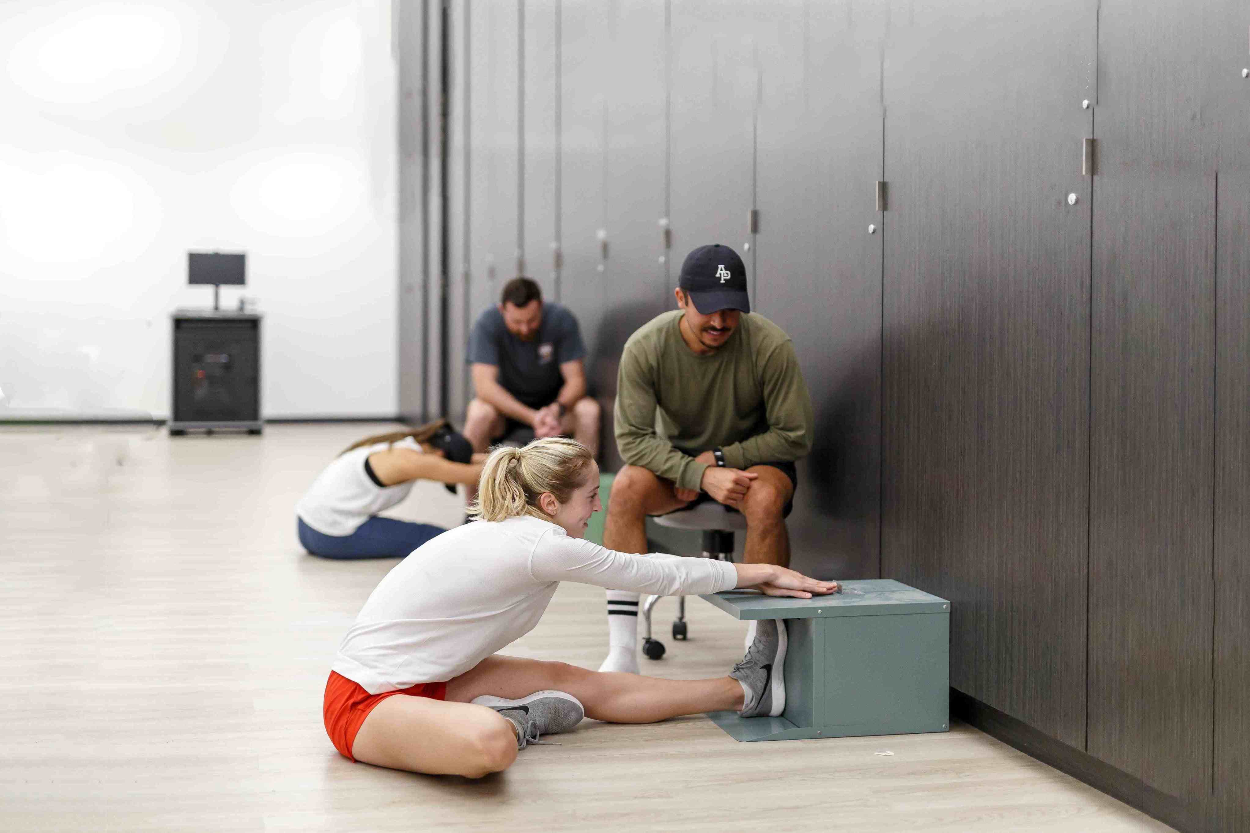 A group of people in an exercise or therapy room, where one person is stretching on the floor while others observe or assist.